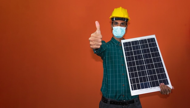 Lavoratore con casco e maschera pandemica che tiene un pannello solare fotovoltaico isolato Lavoratore solare isolato su sfondo arancione