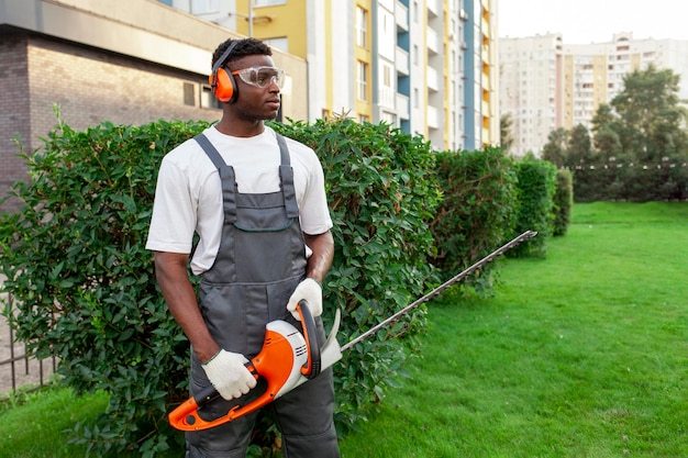 Lavoratore del giardino in uniforme che taglia cespugli uomo afroamericano con occhiali e cuffie