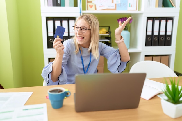 Lavoratore di affari della giovane donna bionda che utilizza computer portatile e carta di credito con l'espressione allegra all'ufficio