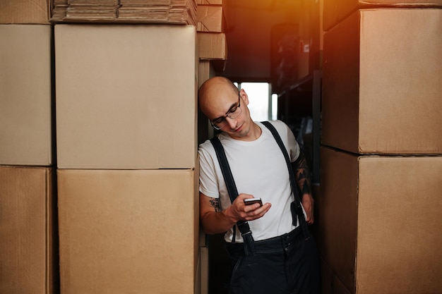 Lavoratore di magazzino maschio stanco che si appoggia su una pila di scatole guardando il suo telefono