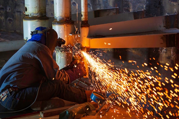 Lavoratore di sesso maschile che utilizza la molatura a scintilla della ruota elettrica sulla conduttura della parte in acciaio al carbonio del metallo della saldatrice all'interno dello spazio confinato del serbatoio