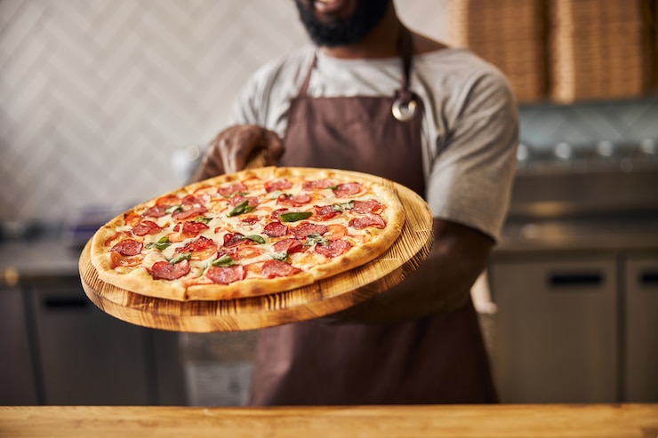 Foto lavoratore di sesso maschile in grembiule che tiene una deliziosa pizza con salame, formaggio e pomodori