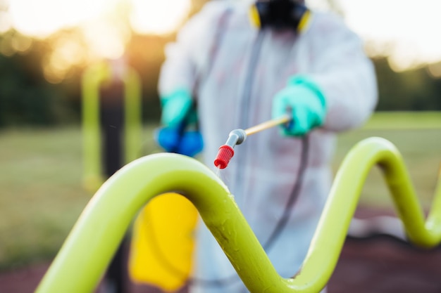 Lavoratore di sesso maschile in prima linea con maschera facciale e abbigliamento da lavoro protettivo che utilizza uno spruzzatore a pressione per disinfettare il parco giochi per bambini. Concetto di protezione e prevenzione della pandemia di virus.