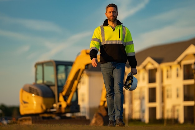 Lavoratore in casco sulla costruzione del sito Lavoratore di bulldozer escavatore uomo Operaio edile con escavatore sullo sfondo Operaio edile con trattore o costruzione in costruzione