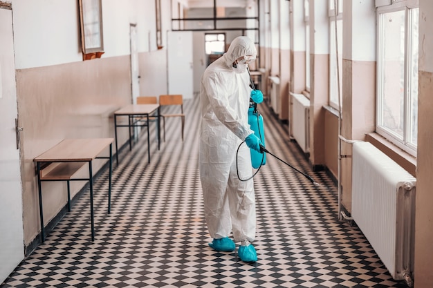 Lavoratore in uniforme bianca sterile, con maschera e occhiali tenendo lo spruzzatore con disinfettante e spruzzando intorno al corridoio a scuola