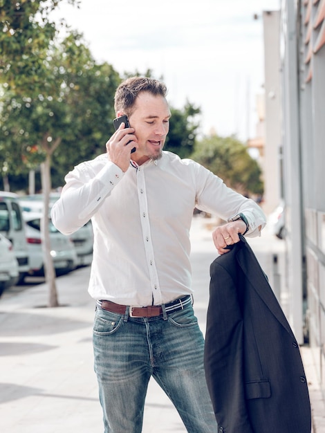 Lavoratore maschio adulto in camicia bianca e jeans che controlla l'ora e parla sullo smartphone prima della riunione di lavoro