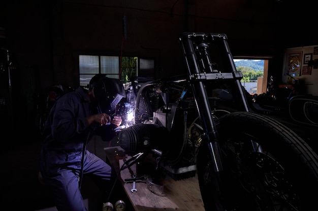 Lavoratore maschio anonimo in uniforme e scudo che utilizza la saldatrice per riparare alcune parti metalliche della bicicletta mentre lavora in officina buia