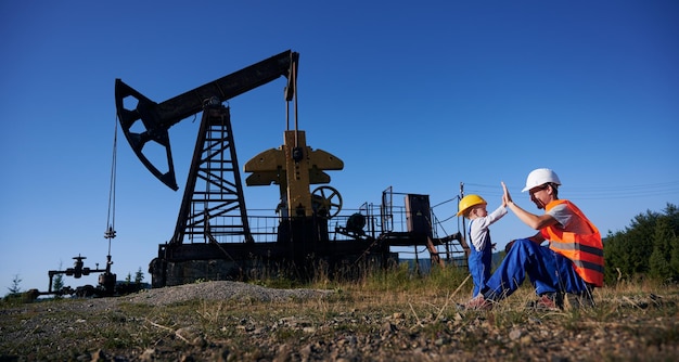 Lavoratore maschio e bambino carino che fanno il gesto del high five mentre trascorrono del tempo nel campo petrolifero con il pompa jack e il cielo blu sullo sfondo L'uomo del petrolio e il bambino adorabile che schiaffeggiano le mani mentre lavorano nel pozzo petrolifero