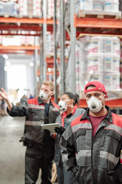 Lavoratore maschio latino di mezza età in uniforme e respiratore protettivo in piedi davanti alla telecamera in magazzino contro i suoi colleghi