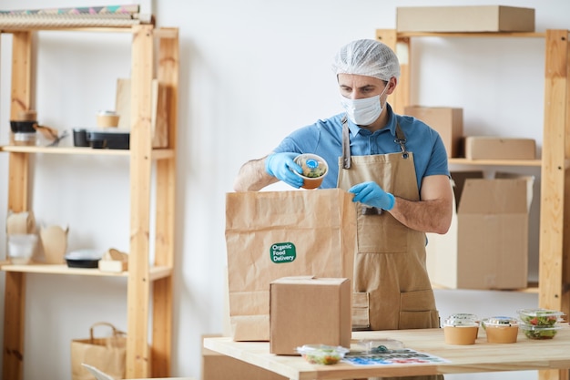 lavoratore maschio maturo che indossa indumenti protettivi mentre impacchetta in modo sicuro gli ordini al tavolo di legno nel servizio di consegna di cibo