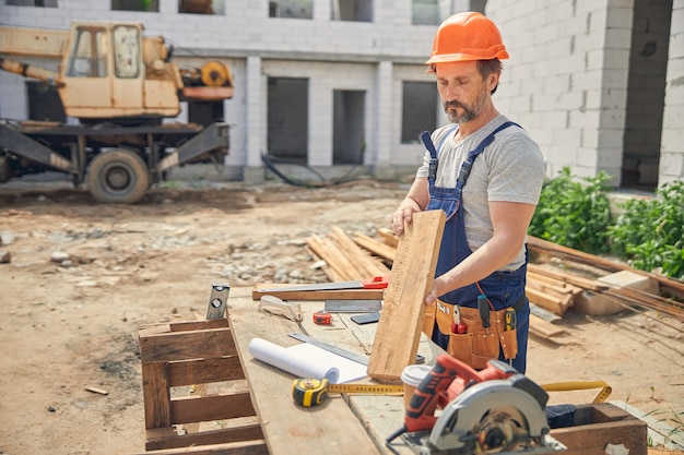 Lavoratore maschio maturo concentrato che fissa una barra di legno nelle sue mani al cantiere