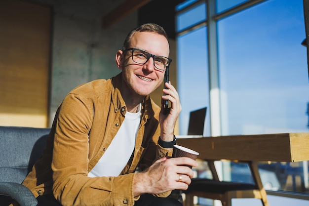 Lavoratore maschio sorridente in abiti casual che distoglie lo sguardo mentre parla al telefono cellulare e beve caffè durante la pausa nel moderno spazio di lavoro creativo