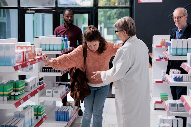 Lavoratore senior della farmacia che aiuta la donna vertiginosa dopo essere svenuta in farmacia mentre diverse persone fanno shopping sanitario acquistando pillole, vitamine. Cliente adulto che gira in negozio accanto agli scaffali