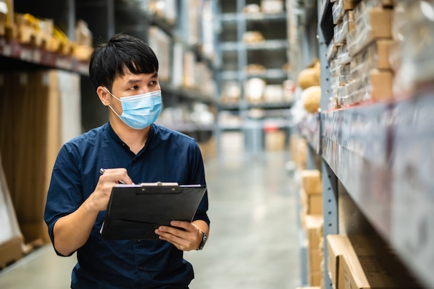 Lavoratore uomo con maschera medica che tiene appunti e controlla l'inventario nel magazzino durante la pandemia di coronavirus