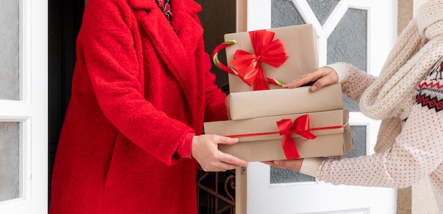 Lavoratore vestito in cappotto rosso che consegna scatole regalo vicino alla porta bianca alla vigilia delle vacanze