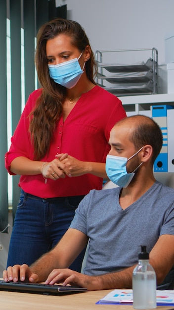 Lavoratori che parlano indossando maschere protettive in ufficio durante il coronavirus. Squadra nel nuovo normale spazio di lavoro dell'ufficio nella società aziendale personale digitando sulla tastiera del computer e puntando al desktop