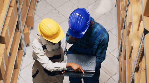 Lavoratori di depositi afroamericani che guardano scaffali con prodotti e scatole di cartone, utilizzando laptop e tablet digitale. Squadra di dipendenti che controllano i pacchi di merci industriali. Tiro a mano libera.