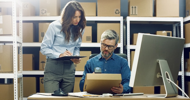 Lavoratrice caucasica della posta che porta scatola all'uomo che lavora al computer nell'ufficio di consegna. Pacco di registrazione maschio e codice a barre di scansione con scanner.