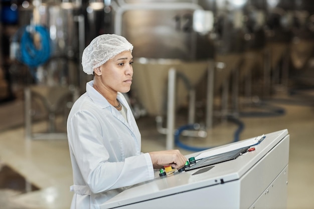 Lavoratrice che indossa camice da laboratorio utilizzando il pannello di controllo presso la fabbrica alimentare