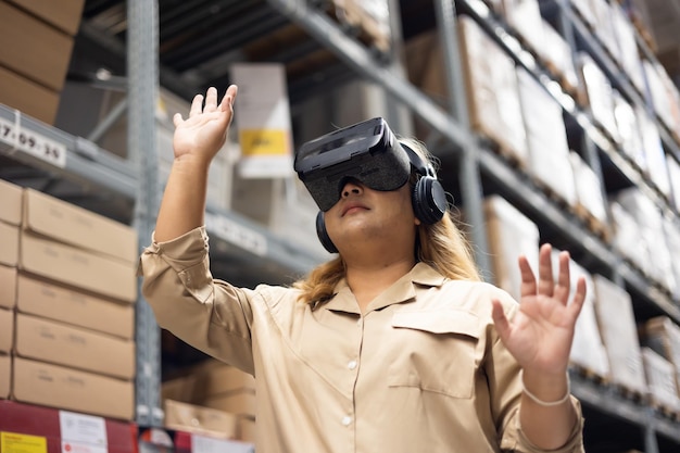 Lavoratrice in plus size che ispeziona scatole di prodotti e utilizza la tecnologia di realtà virtuale per la VR innovativa mentre lavora in un grande magazzino