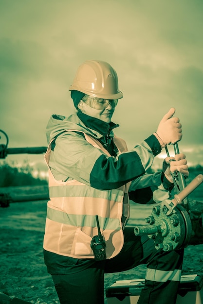 Lavoratrice sul giacimento petrolifero che ripara bene il petrolio e il gas, con la chiave che indossa casco e abiti da lavoro. Processo di lavoro sulla testa del pozzo petrolifero. Seppia tonica.