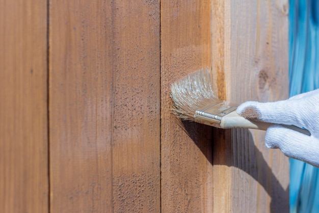 Lavorazione della parete in legno all'esterno. Una mano d'uomo in un guanto dipinge con un pennello le pareti in legno di un gazebo o di una veranda.