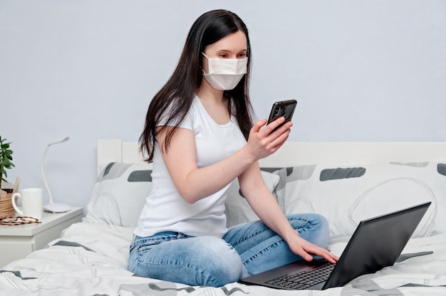 Lavoro a casa o apprendimento online a distanza in quarantena. Donna in maschera protettiva lavorando a distanza sul letto utilizzando il computer portatile e parlando al telefono.