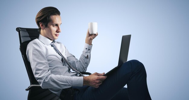 Lavoro a distanza e concetto di freelance con uomo in camicia bianca seduto su una sedia da ufficio con una tazza di caffè in mano e guardando lo schermo del laptop su sfondo azzurro della parete