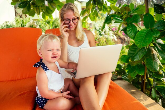 Lavoro a distanza e surf freelance. giovane madre donna d'affari freelance che lavora con il laptop seduto su un lettino accanto alla figlia del bambino. congedo di maternità in una spiaggia estiva di resort tropicale