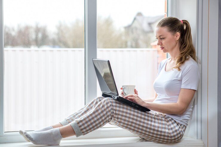 Lavoro a distanza in quarantena domestica. Protezione dal coronavirus. Donna caucasica alla finestra che beve caffè mentre si lavora al computer portatile.