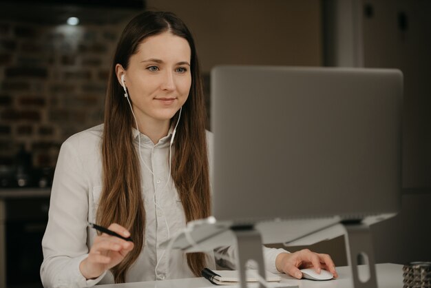 Lavoro a distanza. Una donna caucasica del brunette con le cuffie che lavora a distanza sul suo computer portatile. Una donna d'affari in una camicia bianca fare affari nel suo posto di lavoro a casa.