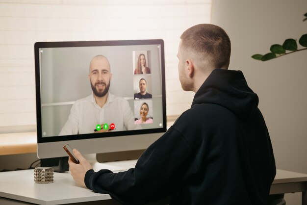 Lavoro a distanza. Una vista posteriore di un uomo durante una videochiamata con i suoi colleghi sul computer desktop. Un collega tiene uno smartphone che lavora da casa durante un briefing online.