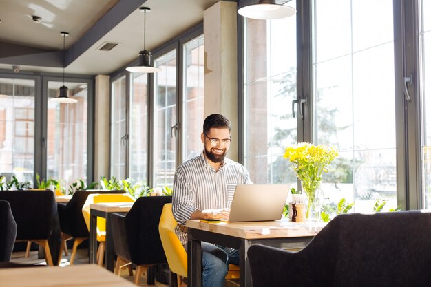 Lavoro a distanza. Uomo felice allegro seduto al computer portatile mentre si lavora dal caffè