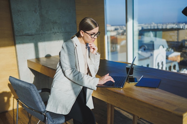 Lavoro d'ufficio attraente giovane manager femminile che lavora in ufficio in piedi in un'area di lavoro luminosa utilizzando un moderno computer portatile giovane donna che beve caffè delizioso