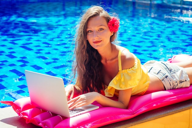 Lavoro da sogno di lavoro a distanza. giovane donna seduta su un materasso rosa gonfiabile in piscina, prendendo il sole e lavorando al computer portatile. ragazza che mangia frutti esotici mango tropicale, ananas e frutta del drago