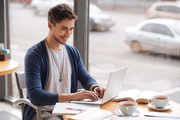 Lavoro freelance. Bel giovane uomo sorridente e lavorando sul computer portatile mentre era seduto al tavolo nella caffetteria.