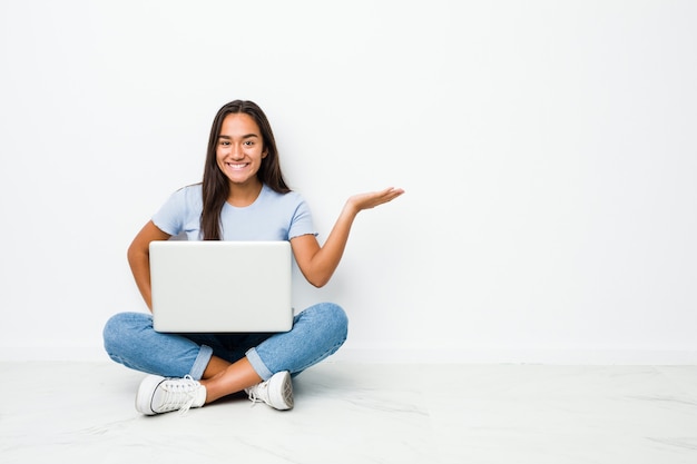 Lavoro indiano di seduta della giovane donna della corsa mista sul computer portatile che mostra uno spazio della copia su una palma e che tiene un'altra mano sulla vita.