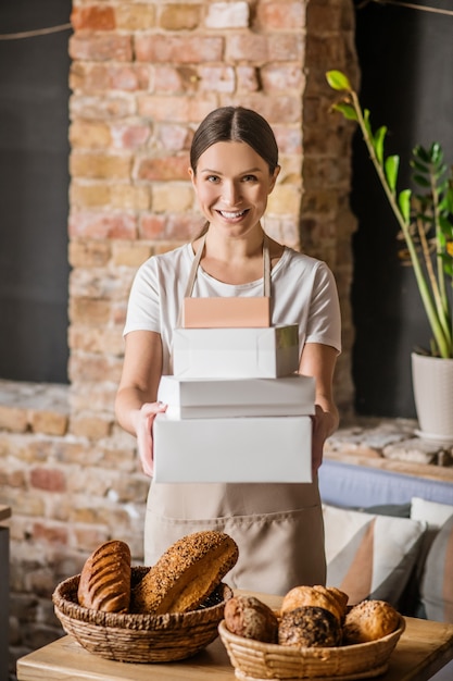 Lavoro preferito. Brillante donna allegra in maglietta leggera e grembiule con scatole vicino al tavolo con pasticcini in panetteria