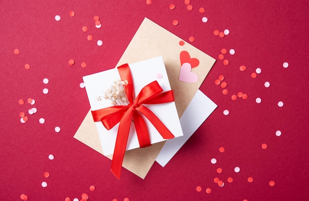 Lay piatto di celebrazione. Confezione regalo bianca e lettera artigianale su sfondo rosso con coriandoli. Concetto di San Valentino, festa della mamma, giorno delle nozze. Vista dall'alto e copia spazio