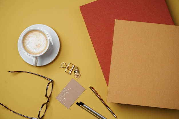 Lay piatto di con libri, tazza di caffè, occhiali da lettura, penne e matite su giallo.