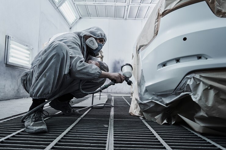 Layanan pengecatan mobil dan perbaikan mobil. Montir mobil dengan pakaian overall putih mengecat mobil dengan pulverizer airbrush di ruang pengecatan
