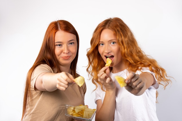 Le amiche dai capelli rossi con una ciotola di vetro stanno tenendo le patatine fritte verso sul muro bianco