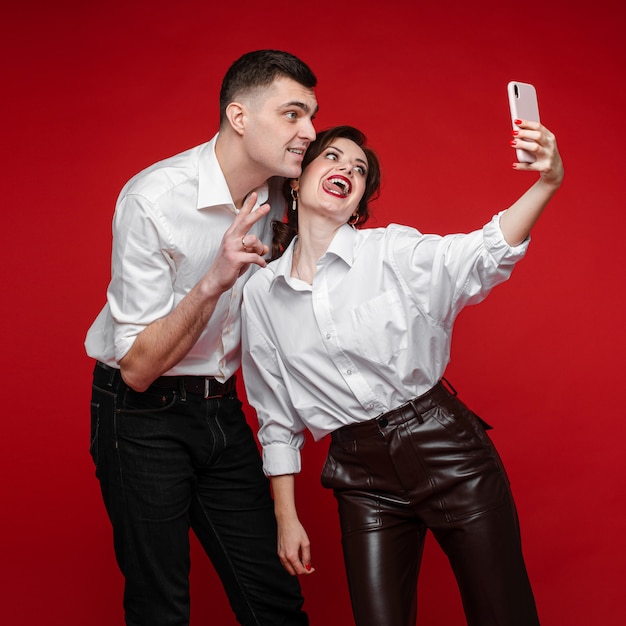 Le belle coppie caucasiche nell'amore prendono il selfie e si divertono molto, immagine isolata sulla parete rossa
