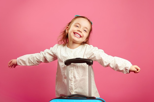 Le braccia estese della bambina sveglia raffigurano un aereo che sorride felicemente si trova vicino alla valigia su un rosa isolato