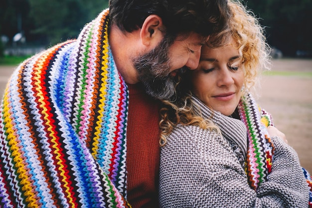 Le coppie adulte romantiche stanno insieme con il sentimento e l'amore coperti da una coperta di lana colorata nel parco all'aperto. Concetto di persone di stile di vita di relazione felice. Tempo libero invernale all'aperto