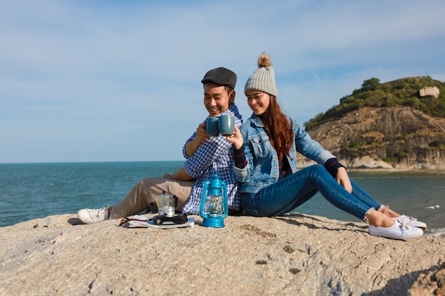 Le coppie asiatiche in camicia blu con la tazza di caffè e la macchina fotografica d'annata fanno un picnic sulla montagna vicino alla vista del mare della spiaggia felice e sorridono affronta