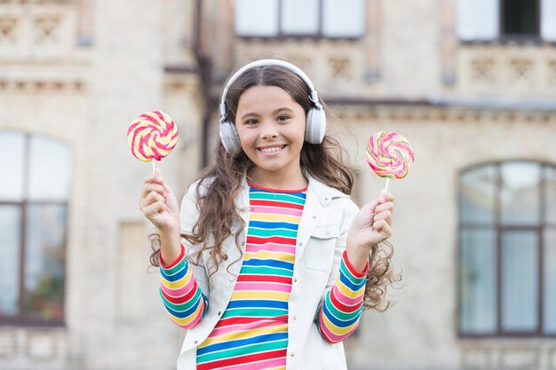 Le cuffie wireless moderne della ragazza allegra mangiano il concetto dell'alimento della via delle caramelle