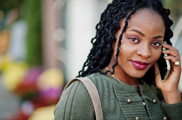 Le donne afroamericane alla moda alla moda in maglione verde e gonna nera hanno posto il caffè all'aperto con la tazza di caffè e parlano sul telefono cellulare.