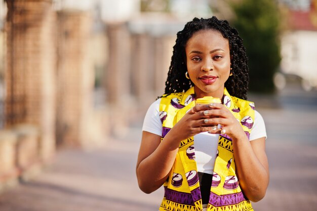 Le donne alla moda dell'afroamericano in rivestimento giallo hanno proposto sulla via con la bevanda calda in tazza di carta eliminabile.