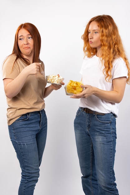 Le donne dai capelli rossi passatempo che indossano magliette e jeans fanno facce su un muro bianco che tiene popcorn